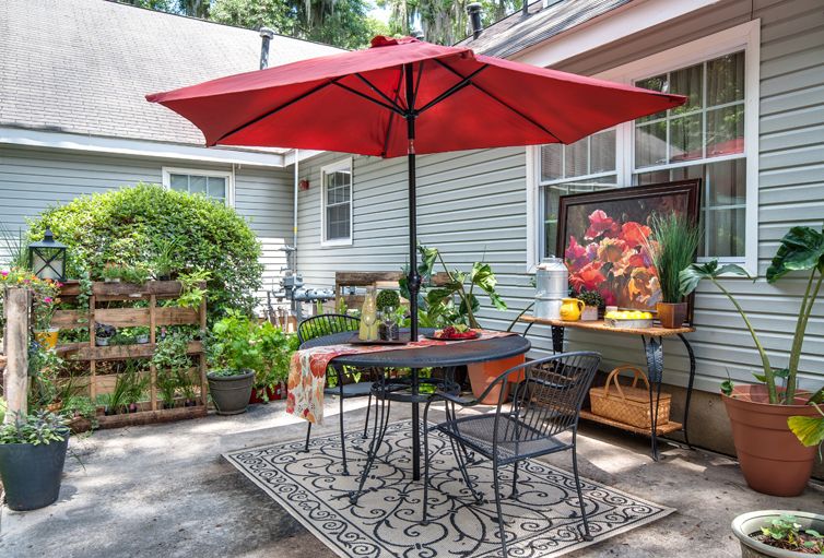 A cozy patio area with a red umbrella, table and chairs, surrounded by lush greenery and potted plants.