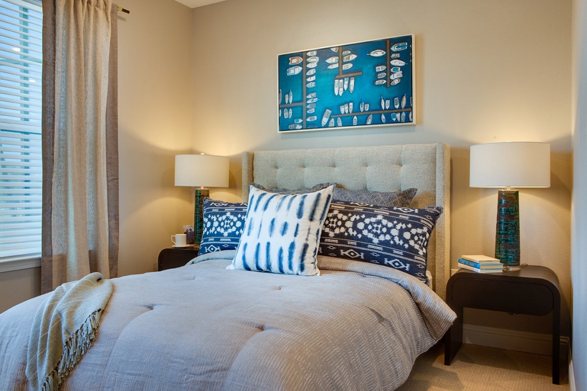 A modern bedroom with elegant decor, featuring a bed with patterned pillows, flanked by lamps on nightstands and artwork on the wall.