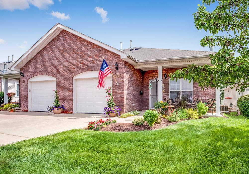 A suburban brick house with a well-maintained lawn, American flag, and two-car garage.