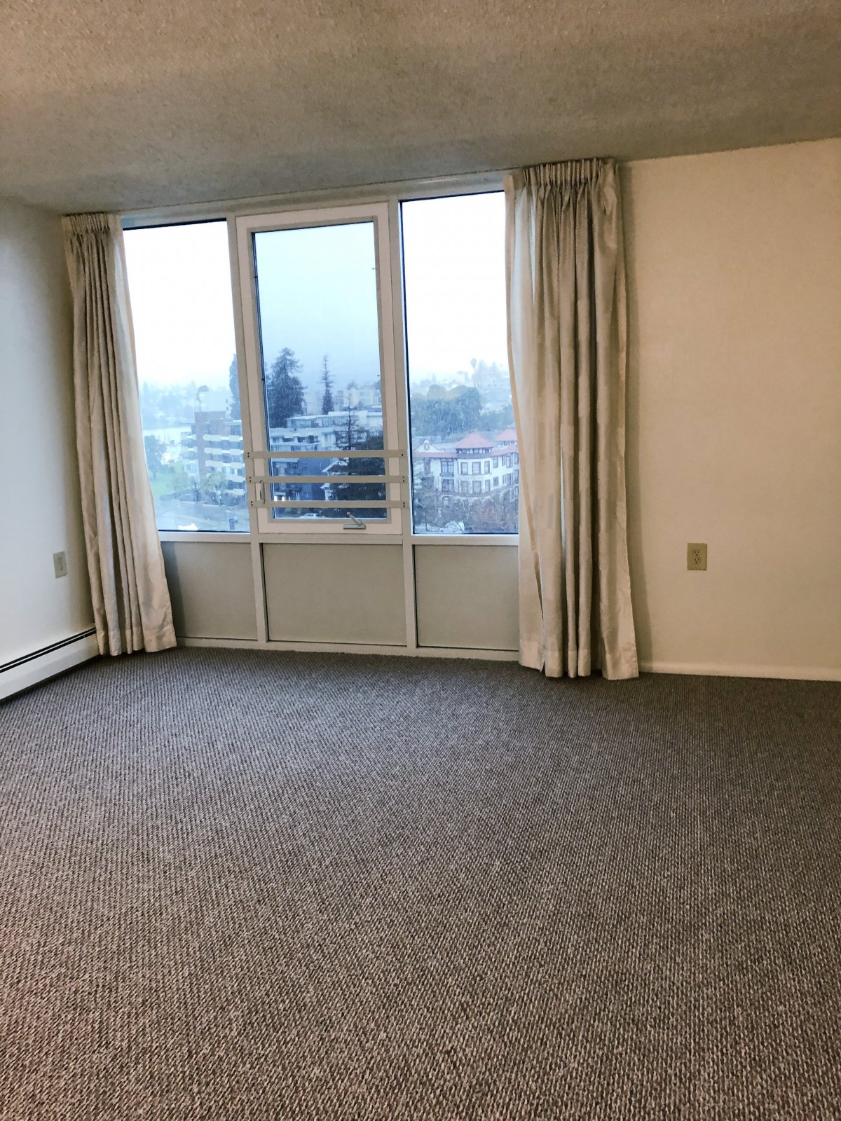 An empty carpeted room with large windows and curtains, overlooking a cityscape.