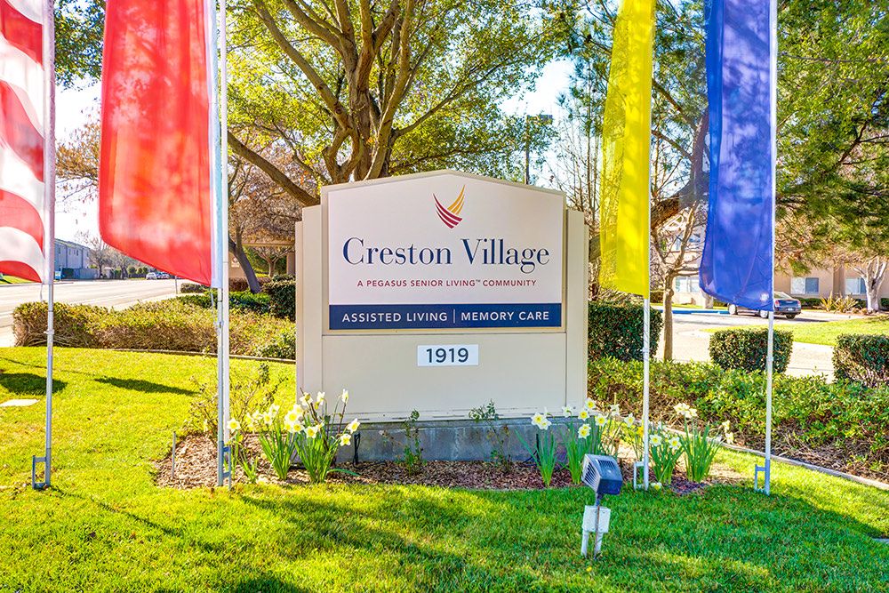 A sign for a retirement community named Creston Village, surrounded by colorful flags and well-maintained landscaping.