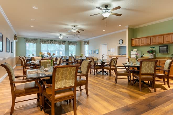 A well-lit dining area with multiple tables and chairs, featuring ceiling fans and large windows.