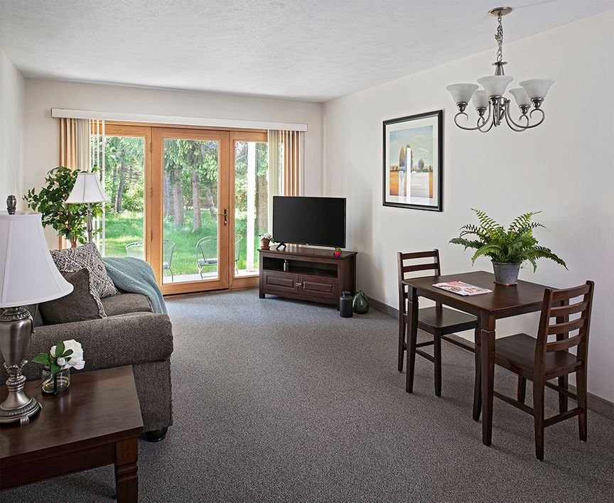 A cozy living and dining area featuring a sofa, television, and a small dining table with chairs, illuminated by natural light from the glass doors.