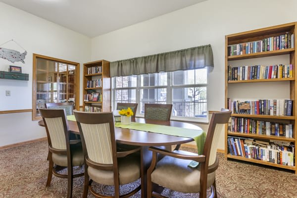 A cozy library or meeting room with a wooden table, surrounded by chairs and bookshelves, lit by natural light from a large window.