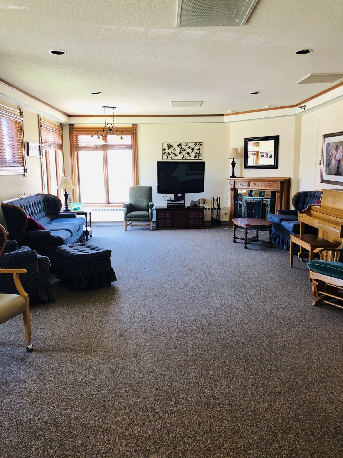 A spacious living room with a television, sofa, piano, and chairs arranged around a central carpeted area.