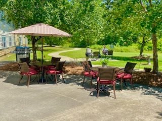 A peaceful outdoor patio with tables, chairs, and an umbrella, surrounded by lush greenery.