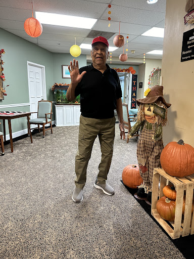 A man cheerfully greets while standing next to festive autumn decorations including a scarecrow and pumpkins.