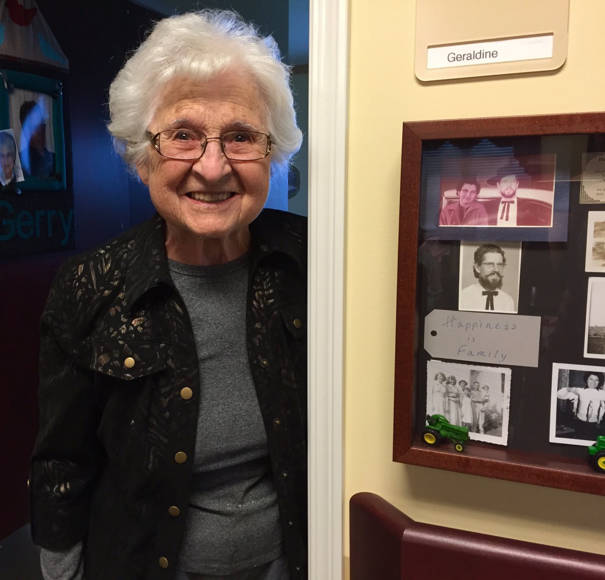 An elderly woman smiling while standing in front of a wall with photographs.