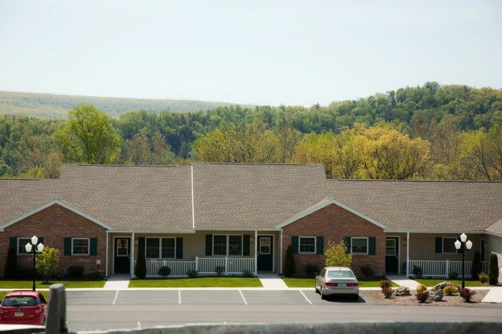 A row of residential buildings with a parking lot in the foreground and a wooded landscape in the background.