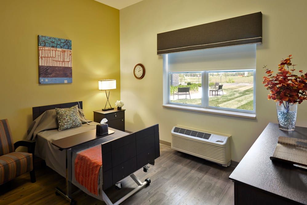 A well-lit hospital room with a single bed, side table, and a large window overlooking green fields.