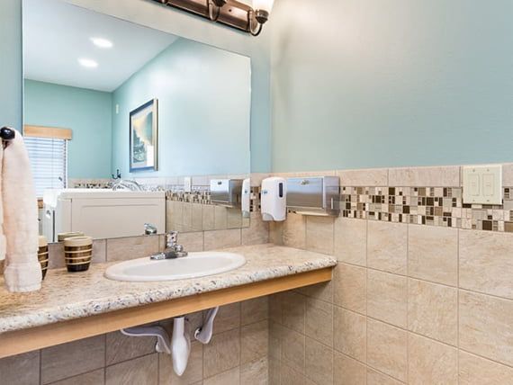 A modern bathroom features a sink with a tiled backsplash, a wall-mounted mirror, and hand soap dispensers.