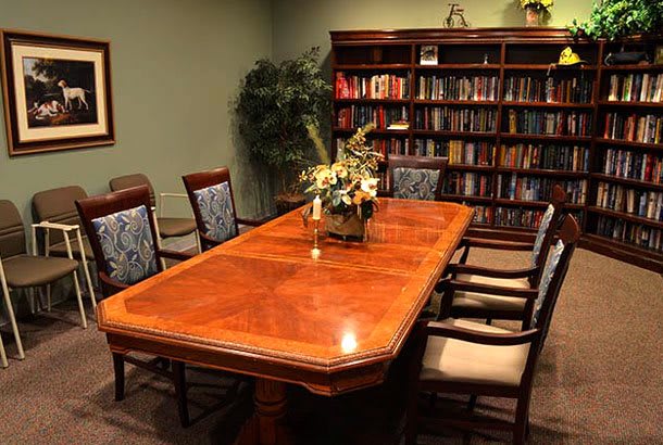 A conference room with a polished wooden meeting table surrounded by chairs, a bookshelf in the background, and decorative plants.
