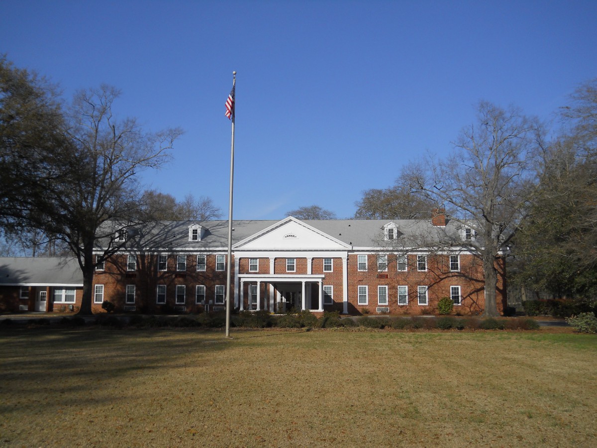 A red brick building with a flagpole in front, surrounded by trees and grass on a sunny day.