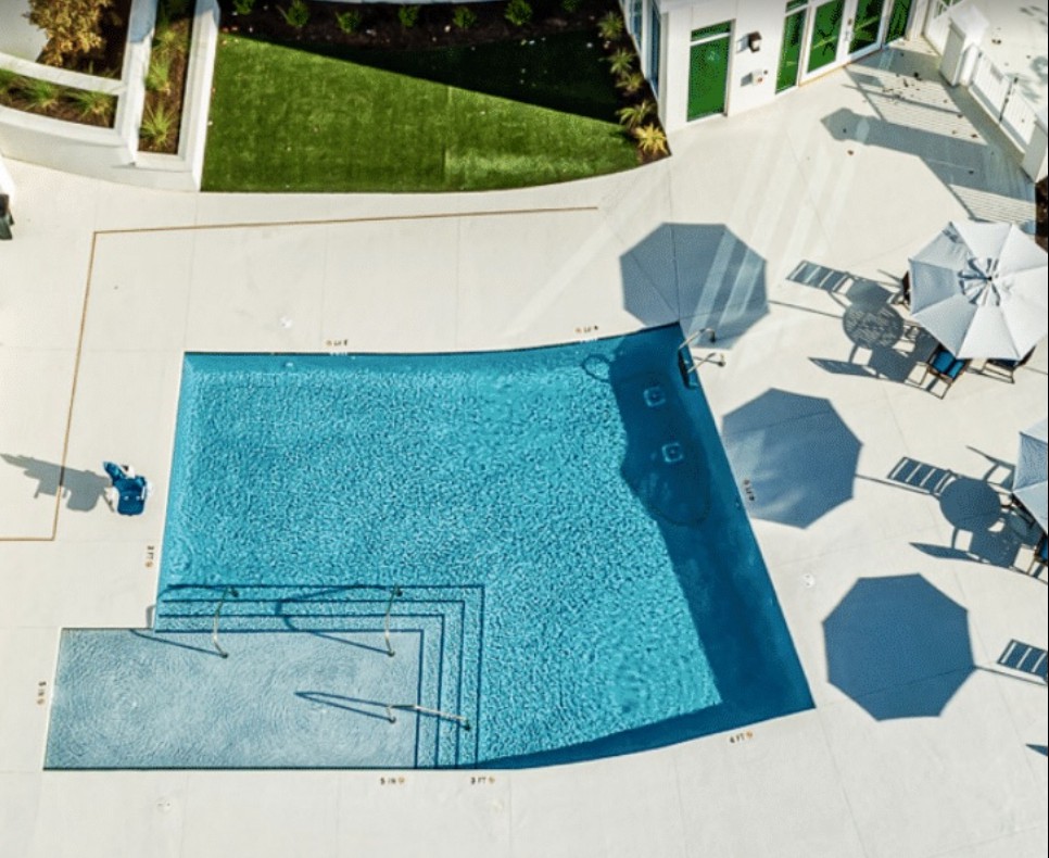 An aerial view of a swimming pool surrounded by patio furniture and umbrellas on a sunny day.