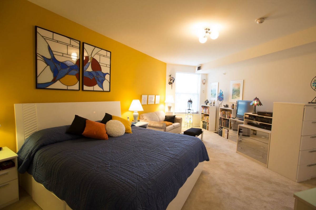 A modern, cozy bedroom featuring a yellow accent wall, artwork above the bed, and comfortable furniture.
