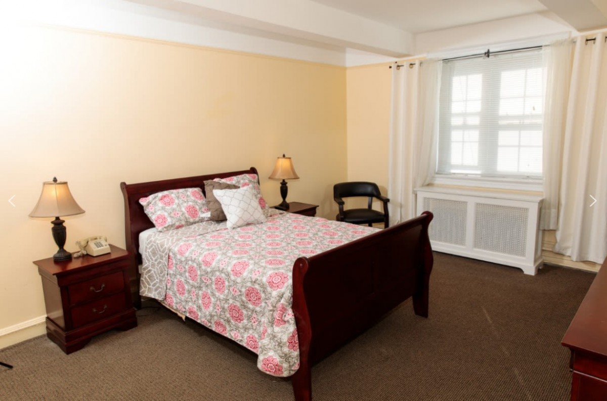 A cozy bedroom with a neatly made bed, a lamp on a nightstand, and a chair near the window.