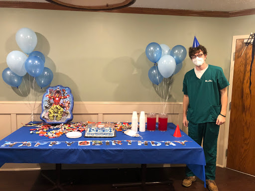 A festive birthday setup featuring blue decorations, a cake, and a person in medical scrubs celebrating.