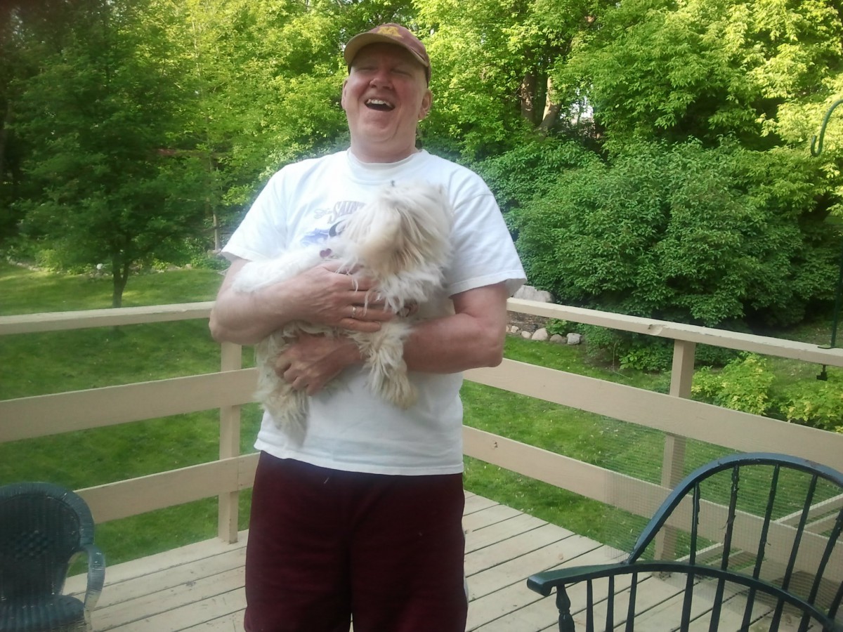A person smiling and holding a dog on a wooden deck surrounded by greenery.