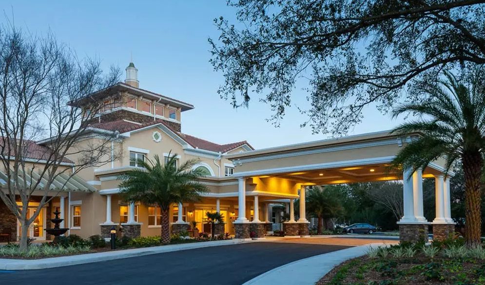 A well-lit building entrance with palm trees and a driveway at dusk.