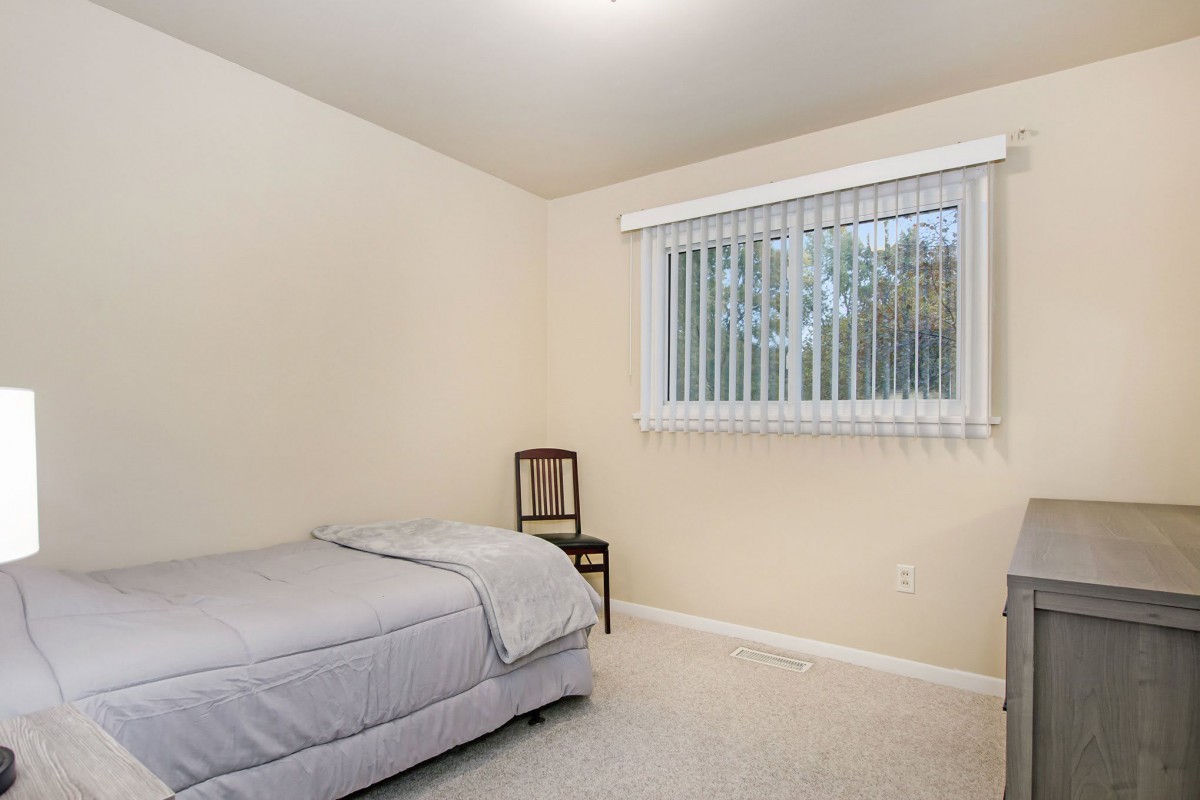 A minimalist bedroom with a single bed, chair, and window with blinds.