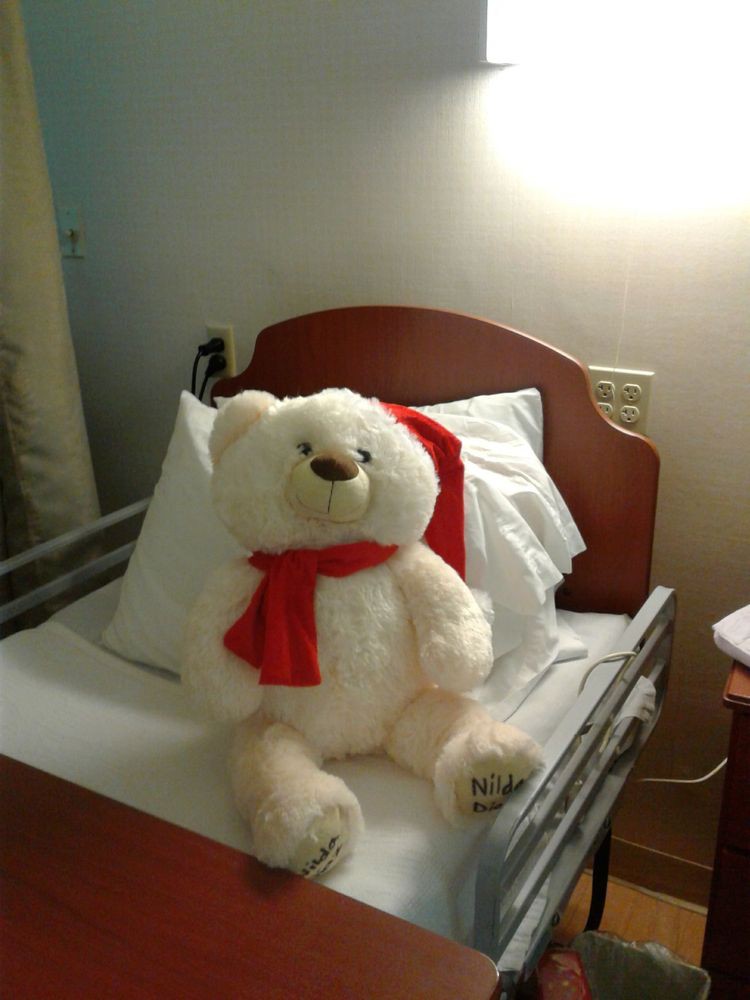 A teddy bear with a red scarf sitting on a bed in a hospital room.