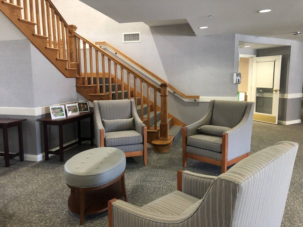 A cozy lobby area featuring comfortable chairs, an ottoman, and a wooden staircase.