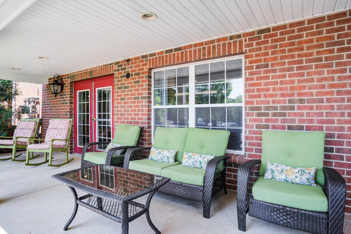 A cozy porch featuring comfortable outdoor seating with green cushions, set against a brick wall and glass doors.