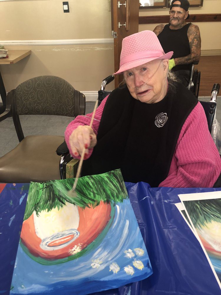 An elderly woman wearing a pink hat is painting a picture at a table.