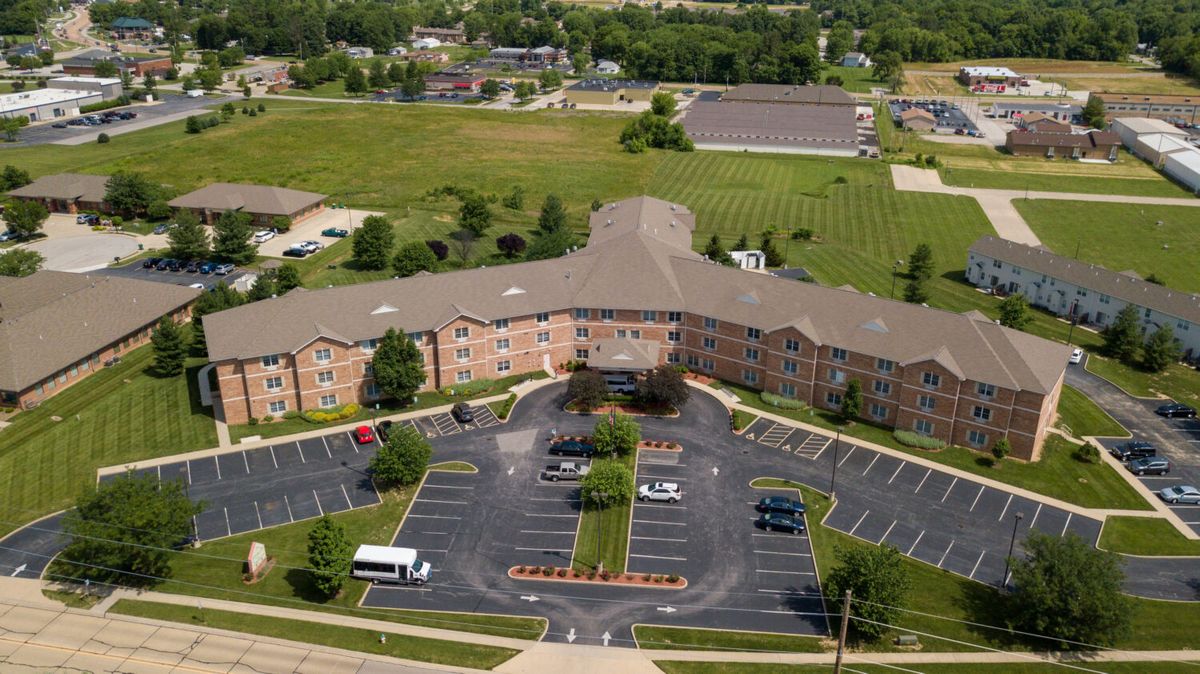 Aerial view of a large, L-shaped building with an adjacent parking lot and surrounding greenery in a suburban setting.