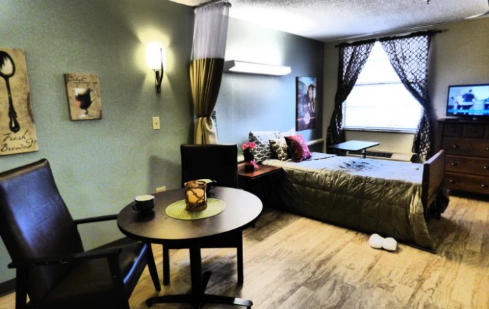 A cozy studio apartment with a comfortable bed, small dining table, and ample natural light from the window.