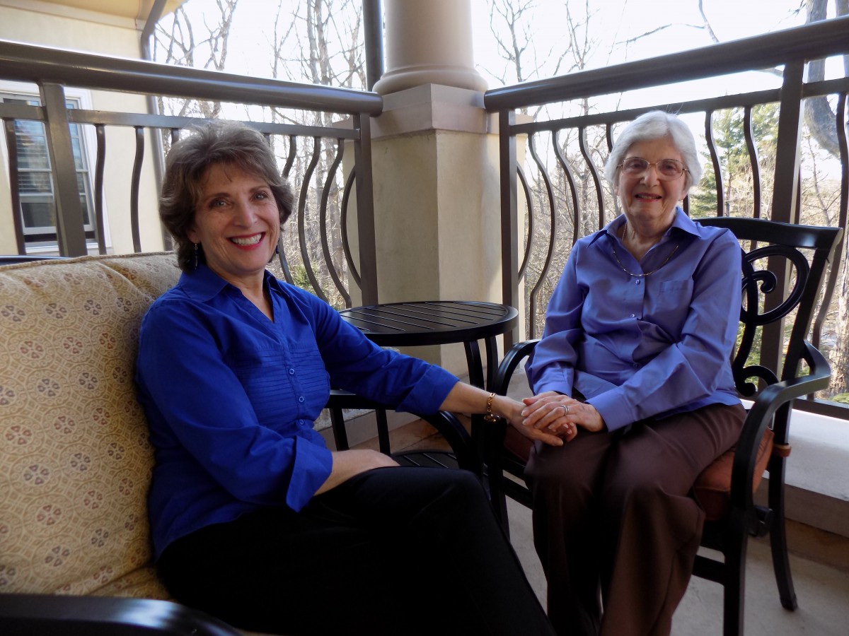 Two women sitting on a balcony, holding hands and smiling.