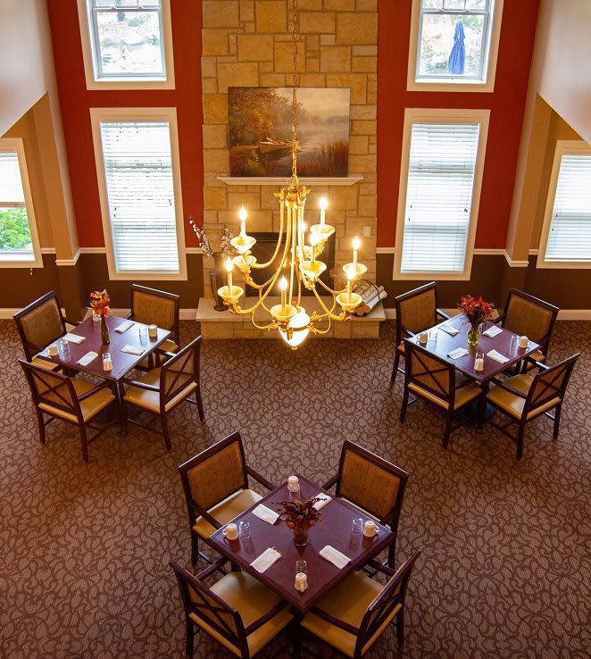 A well-lit dining room featuring a grand chandelier, with neatly arranged tables and chairs on a carpeted floor.