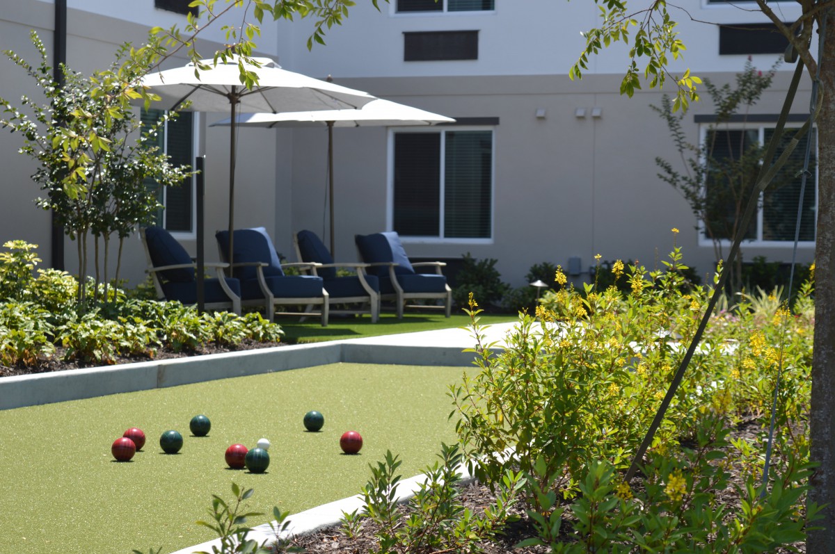 A serene outdoor space featuring a bocce court surrounded by lush greenery, complete with lounge chairs and umbrellas for relaxation.