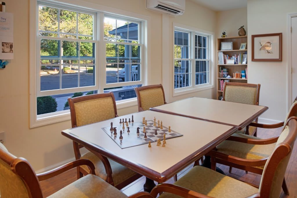 A cozy game room featuring a chess set arranged on a table, surrounded by comfortable chairs and ample natural light from large windows.