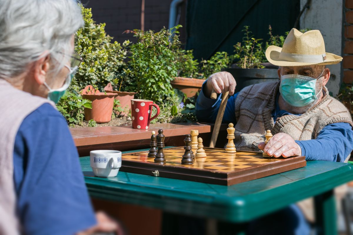 Two elderly individuals playing chess outdoors while wearing masks.