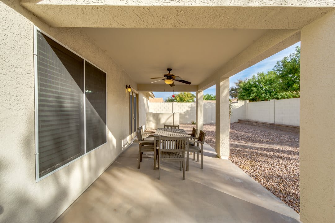 A peaceful covered patio with a dining table and ceiling fan, overlooking a sunny backyard.