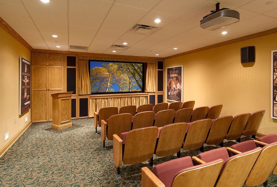 A cozy home theater setup with rows of seating and a large screen, featuring wood paneling and a carpeted floor.