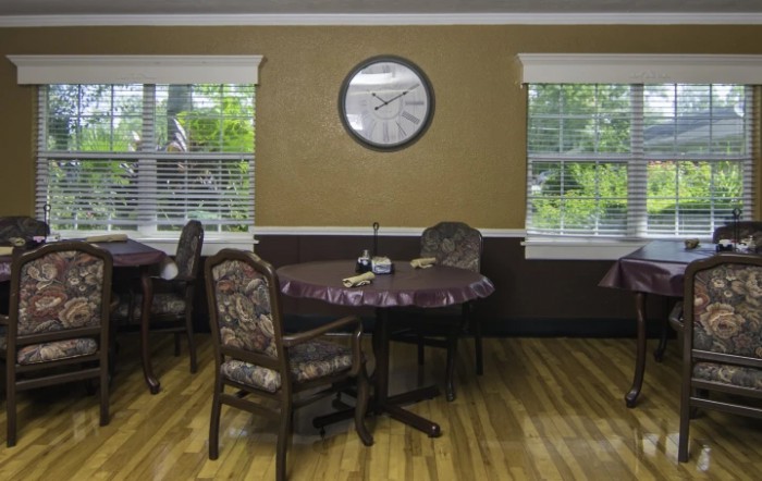 A cozy dining room with tables and chairs, featuring a clock on the wall and windows overlooking greenery.