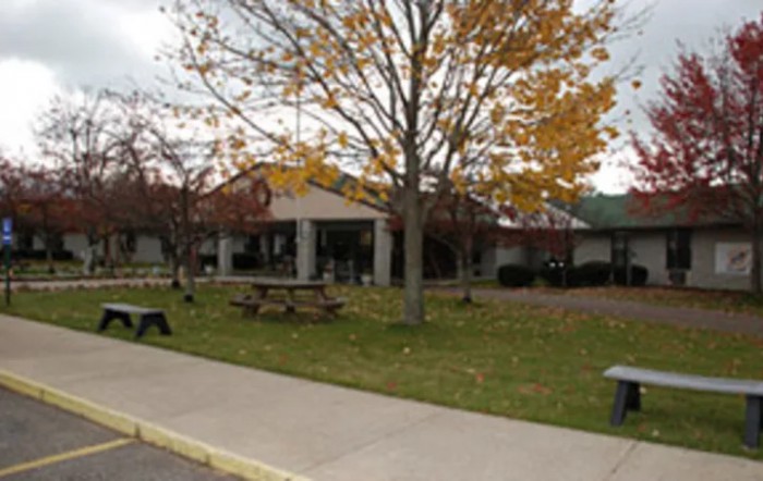 A view of a building surrounded by trees with autumn foliage, featuring picnic tables and a sidewalk.
