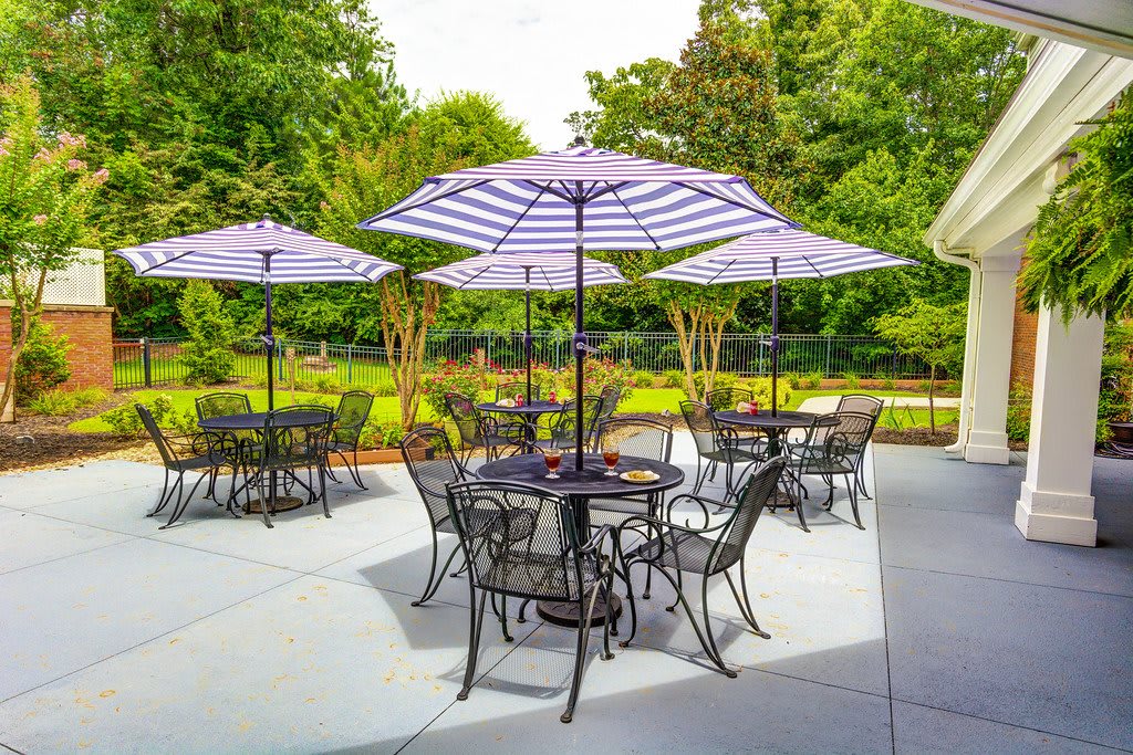A sunny outdoor patio with tables and chairs under striped umbrellas, surrounded by lush greenery.