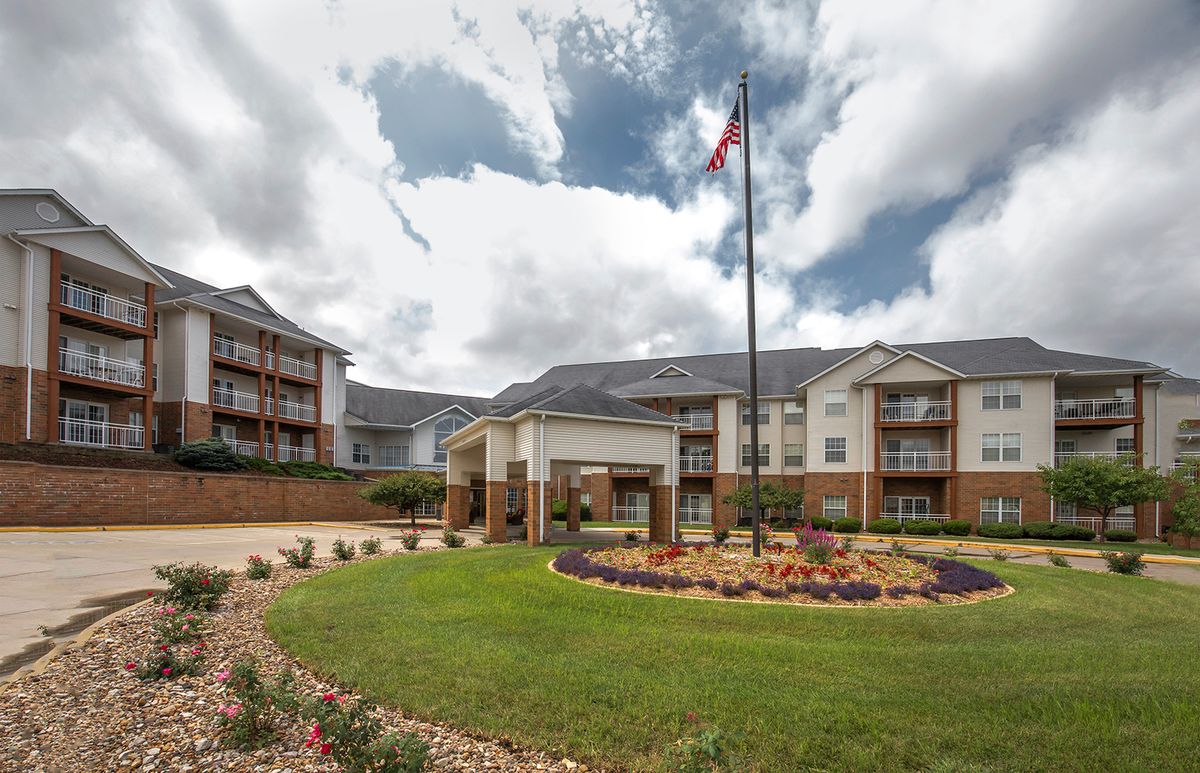 A multi-story apartment building with a landscaped garden and flagpole at the entrance under a cloudy sky.
