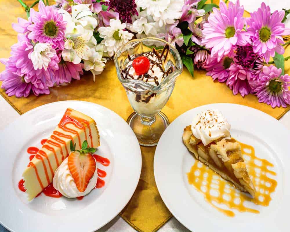 A tempting array of desserts featuring a strawberry-topped cake, a scoop of ice cream with caramel sauce, and a chocolate-drizzled sundae, set against a backdrop of colorful flowers.