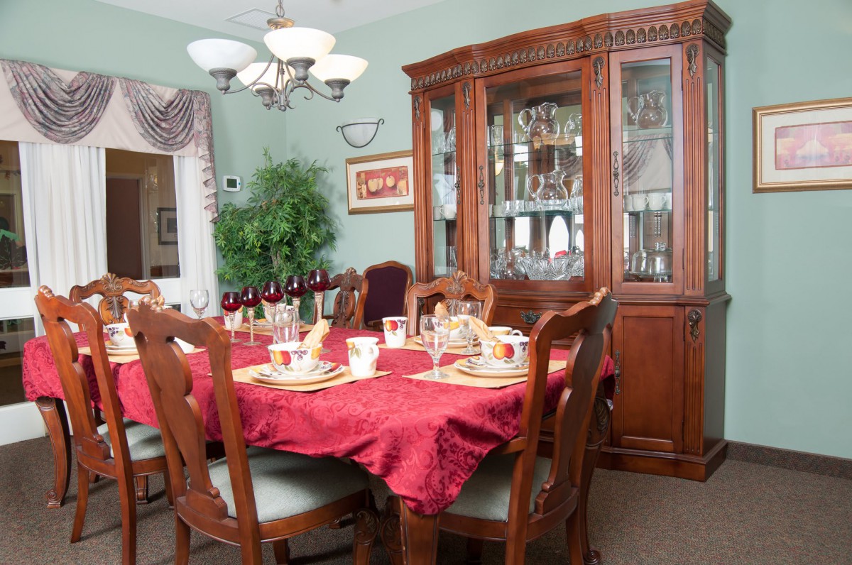 An elegant dining room with a wooden table set for a meal, featuring a red tablecloth and surrounded by matching chairs. A china cabinet is in the background, adding to the sophisticated decor.