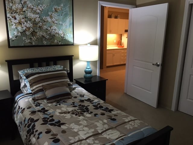 A cozy bedroom with a bed, nightstand, and lamp, leading to an open door with a view of the adjacent room.