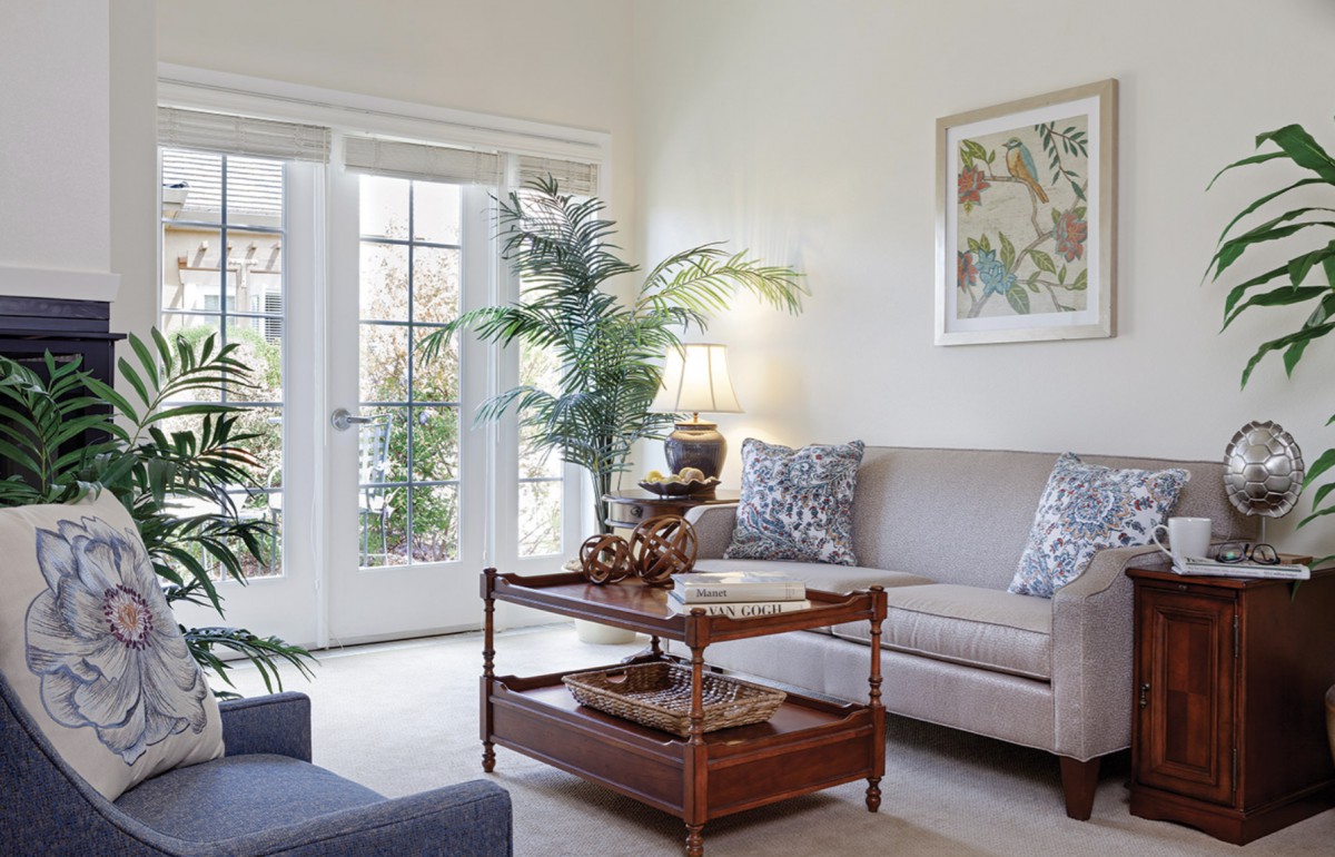 A cozy living room with neutral tones, featuring a sofa, a wooden coffee table, and elegant decor, bathed in natural light from large French doors.