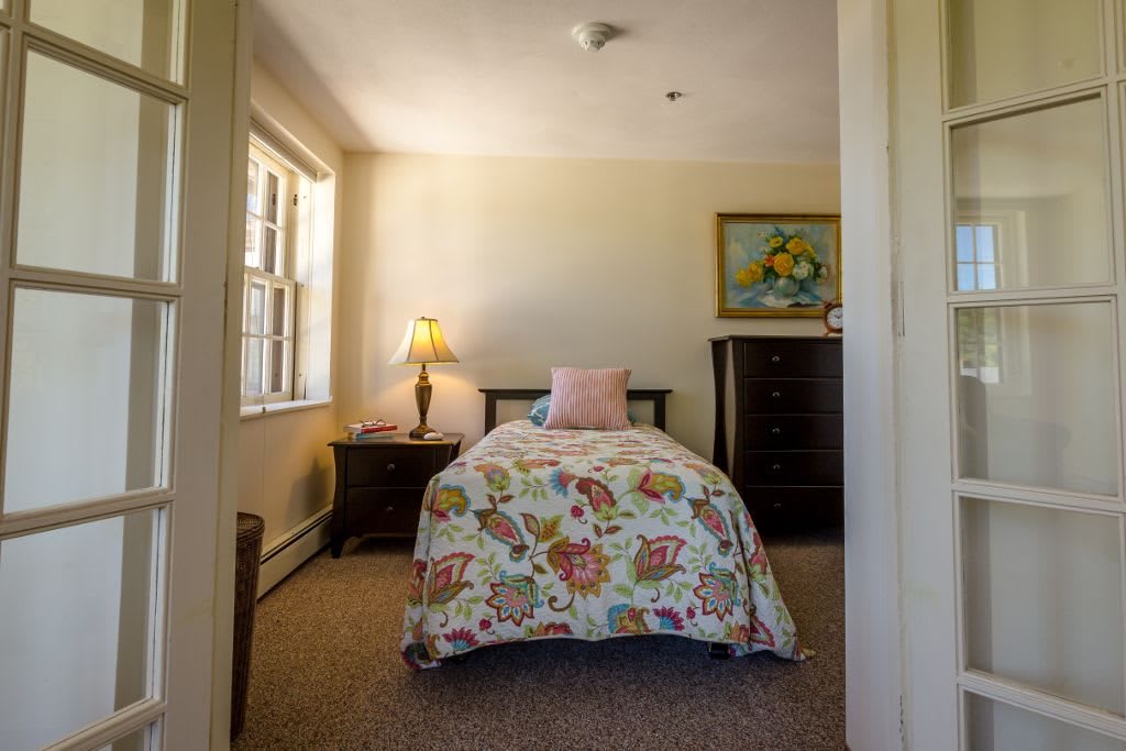 A cozy bedroom with a single bed, floral bedding, and soft lighting from a lamp by the window.