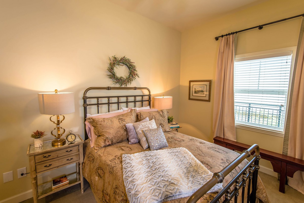A cozy, well-decorated bedroom featuring a comfortable bed with pillows and throws, two lamps on side tables, and a window with curtains.