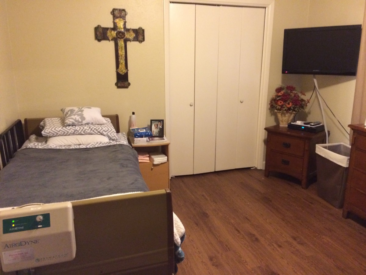 A small bedroom with a hospital bed, crucifix on the wall, TV, and a wooden side table.