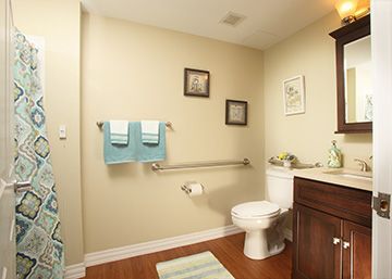 A well-lit bathroom with a toilet, wooden floor, and accessible handrail. Towels hang on the wall alongside framed pictures.