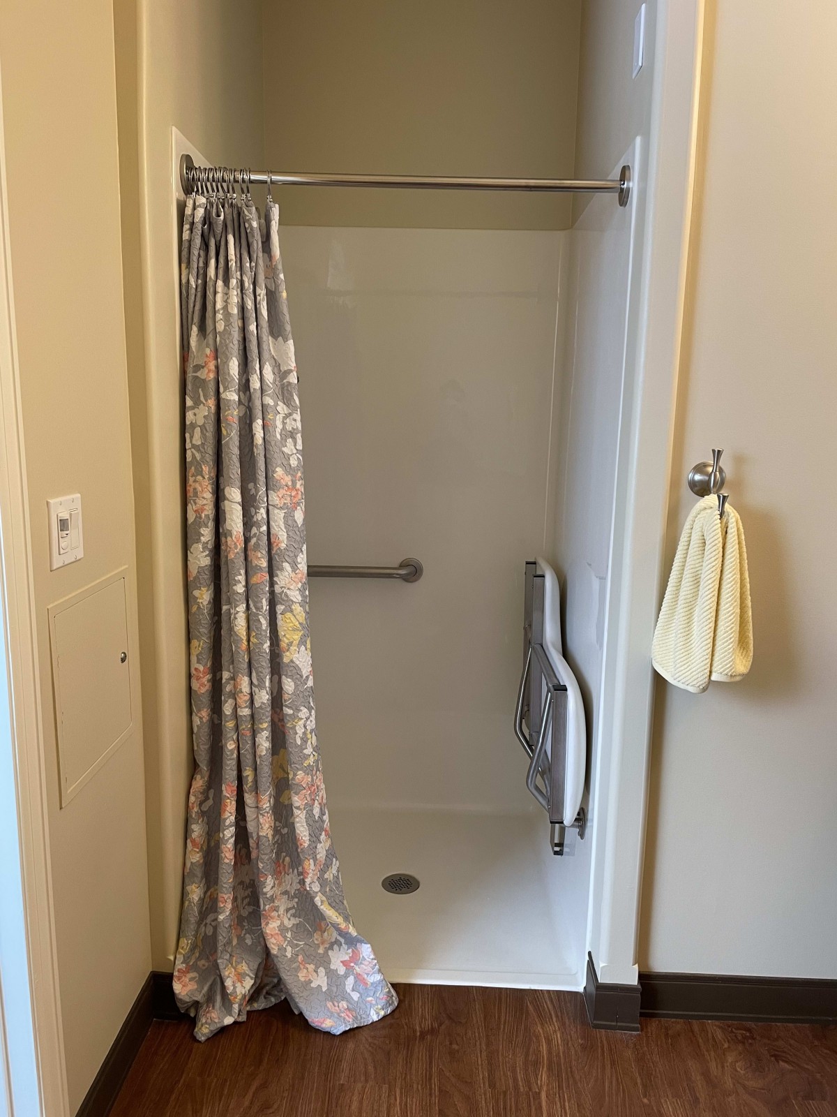 A bathroom with a shower stall, floral curtain, handrails, and a towel hanging on the wall.